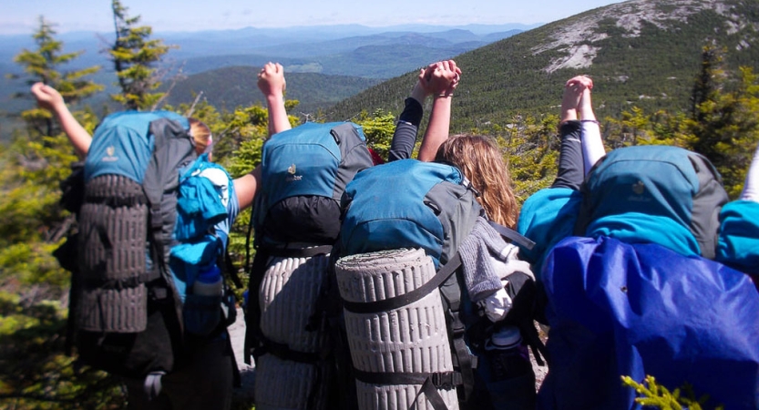 A group of students wearing backpacks hold hands and raise them in celebration above a mountainous landscape. 